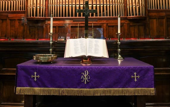 Church altar with purple Chi-Rho cloth, open Bible, gold offering plate, and candles